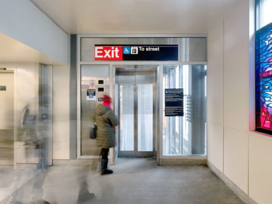 A woman wearing a winter coat and hat waits for elevator with a glass door at subway station.