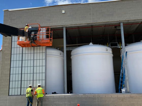 Three men in safety gear work at a large building opening with a translucent doorway. One man is on a lift at the top of the open and the others work at below the lift.