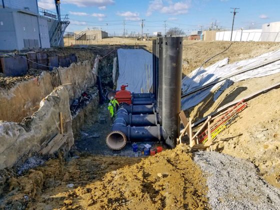 Construction workers in safety gear work beside large, connected pipes in a deeply excavated opening.