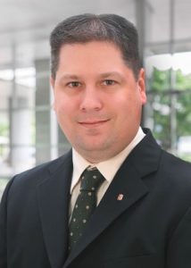 A man is posing for a professional headshot wearing a black suit jacket, white shirt, and dark green tie.