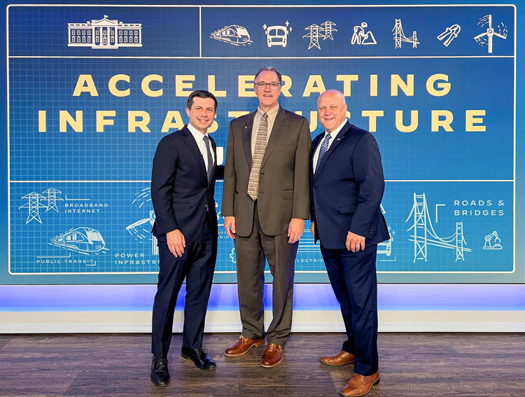 Three men standing next to each other in front of a blue sign smiling dressed in suits and ties.