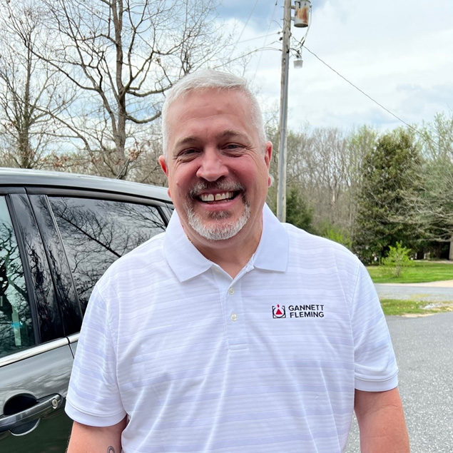 A man with gray hair and a beard wearing a white shirt smiling for the camera.