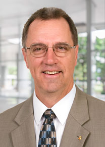 A man in a tan suit and glasses smiling to the camera.