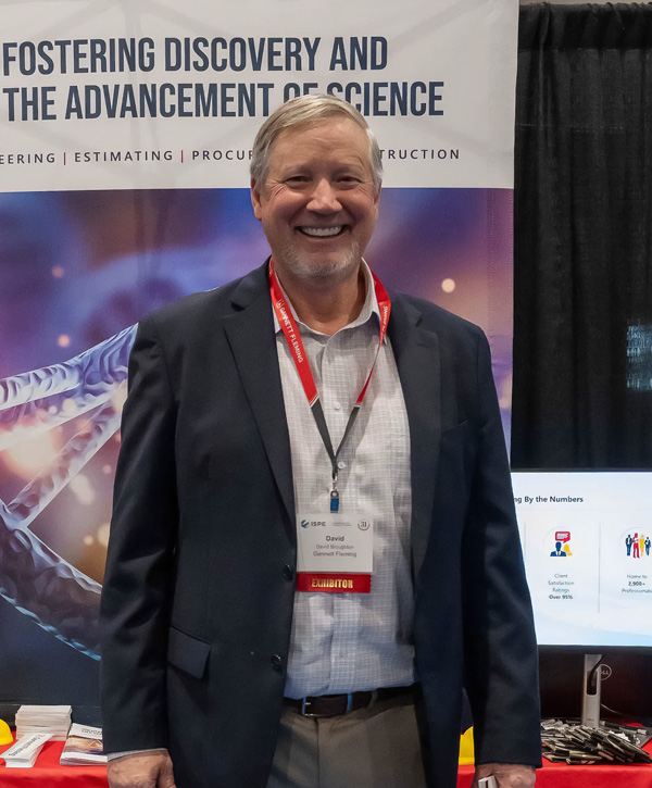 A cheerful man, wearing a navy blazer, checked shirt, and red lanyard, stands in front of a Gannett Fleming exhibit booth. The booth features a red tablecloth with promotional items, a banner reading “Fostering Discovery and the Advancement of Science,” and a DNA helix image.