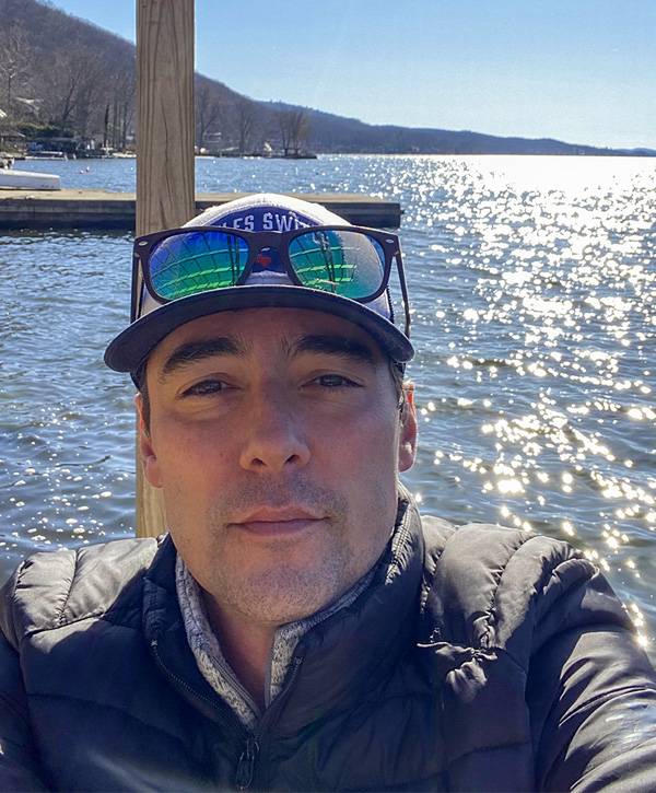 Bryan is sitting on a dock enjoying a beautiful, sunny day on the lake. He is wearing a hat, sunglasses on his head, and a black puffy jacket.