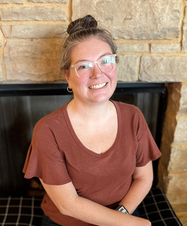 Chelsea is sitting on her hearth on a plaid blanket, smiling. She is wearing glasses, a brown shirt, jeans, and her hair in a bun.