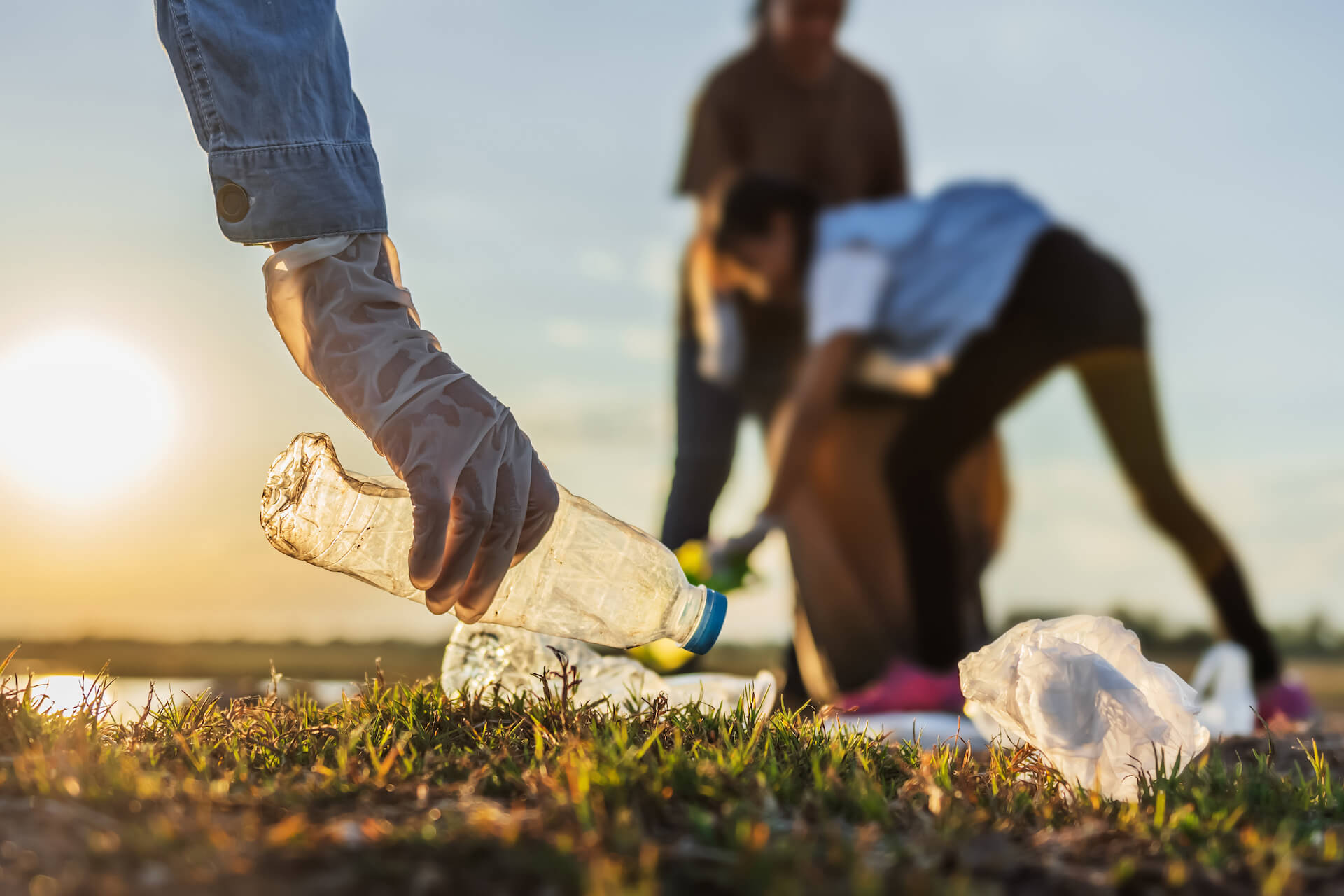 1. A gloved hand picks up a plastic water bottle from a grassy yard while two people in the background also pick up trash.