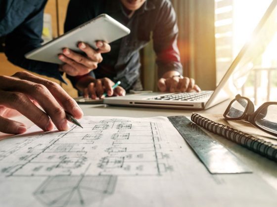 Two people at a worktable. The person in the back is standing over a laptop, while the other person has a pencil over project renderings.