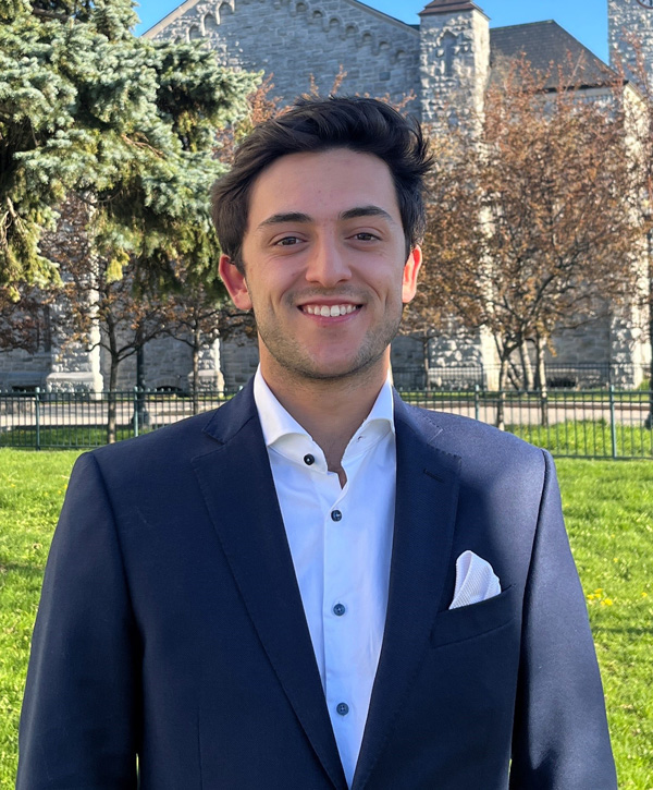 A man in a blue suit and white shirt is smiling in front of a stone building.