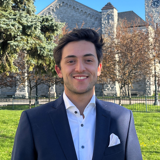 A man in a blue suit and white shirt is smiling in front of a stone building.