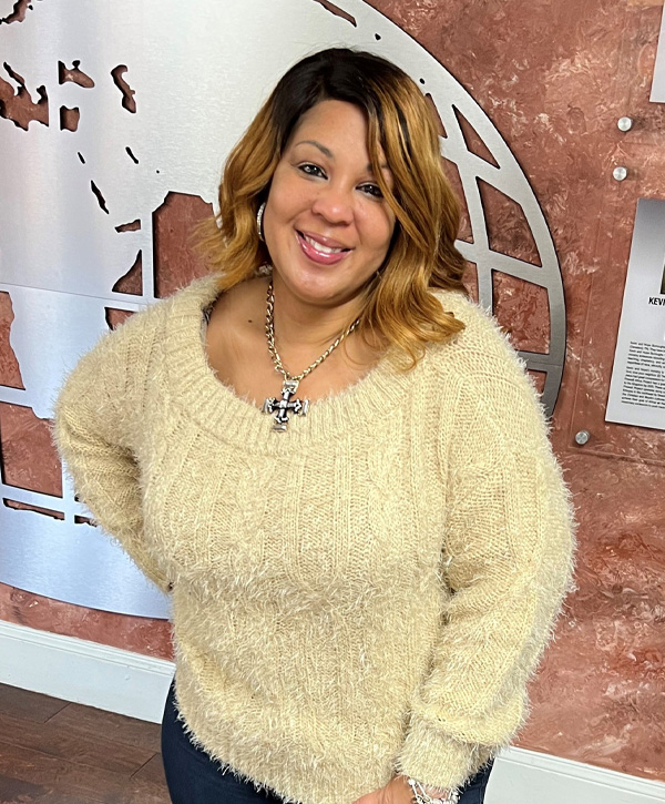 Cybersecurity expert Tamika Bass smiles at the camera with her right hand on her hip while wearing a tan sweater in front of a brown wall.