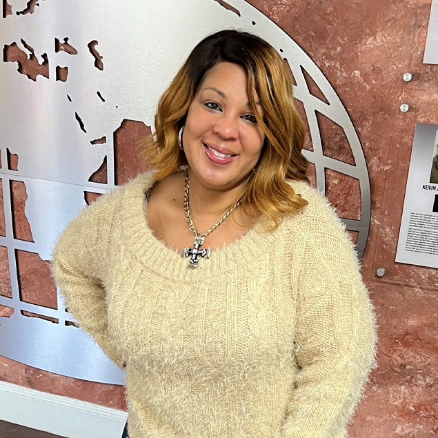 Cybersecurity expert Tamika Bass smiles at the camera with her right hand on her hip while wearing a tan sweater in front of a brown wall.