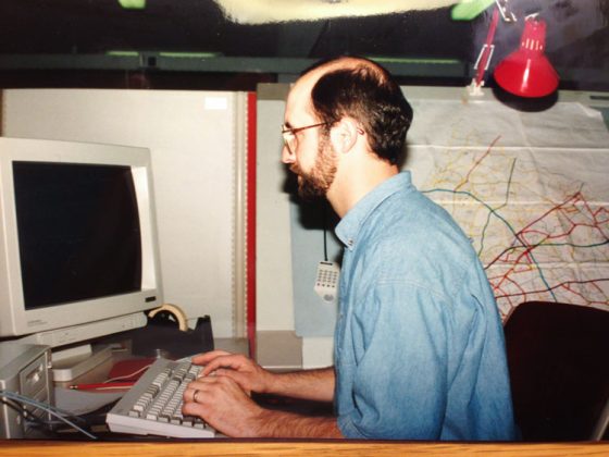 Dave working in 1994. Wearing a blue button-down shirt, he sits at his desk using a desktop computer with a map behind him.