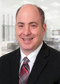 A male in a dark suite with a white shirt and red tie smiling.