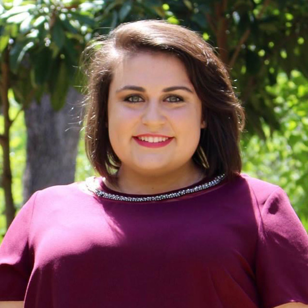 HR Professional Laura Dorval stands outside smiling on a sunny day and wearing a maroon short-sleeved top with lots of greenery in the background.