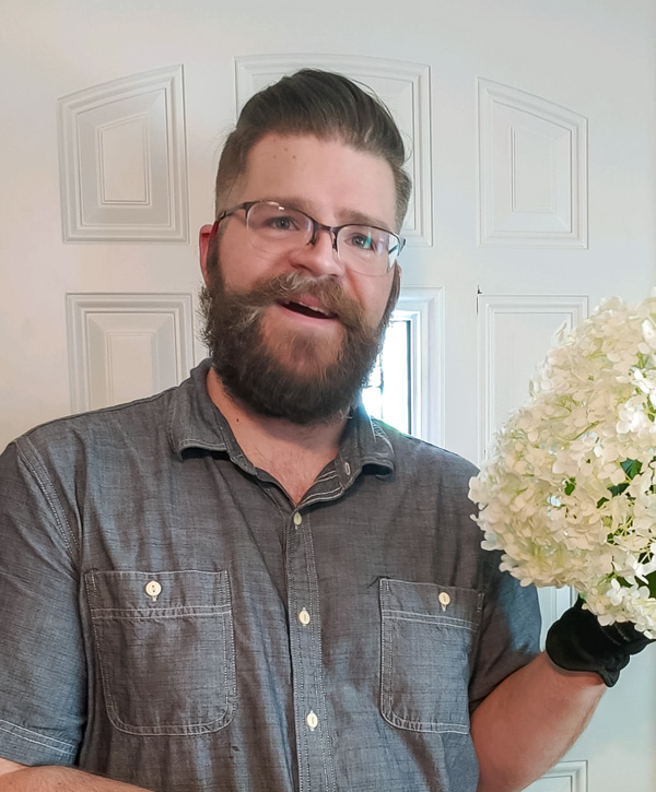 Jesse is standing in his doorway holding a bunch of white flowers he picked from his garden. He is wearing a gray shirt and black gloves.