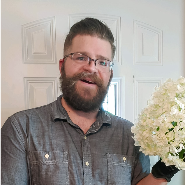 Jesse is standing in his doorway holding a bunch of white flowers he picked from his garden. He is wearing a gray shirt and black gloves.