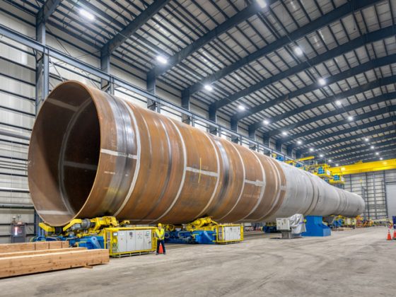 A steel monopile in EEW Group’s manufacturing facility. The monopile is 40 feet wide and 400 feet long. A man standing next to the monopile is small in comparison.