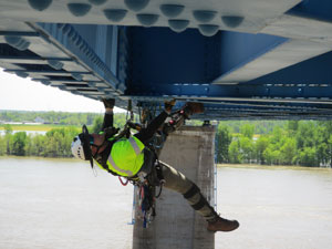 A man in a high-visibility vest hanging upside down while inspecting the underside of a bridge using rope to secure himself.