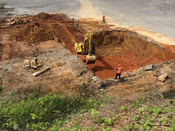 Am aerial view showing two construction workers in PPE working alongside heavy machinery as it digs a large hole.