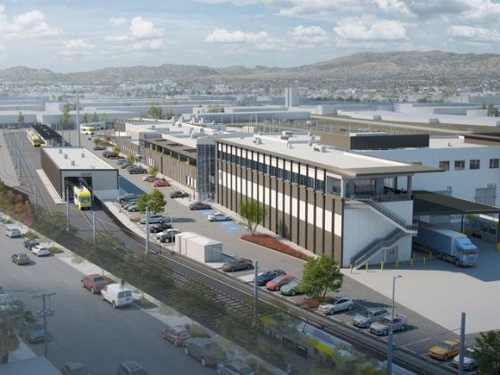 Electric rail cars entering East San Fernando Valley LRT maintenance facility, mountains in the background, cars parked on street adjacent to facility