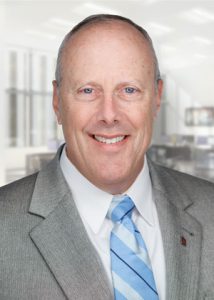 A man is posing for his headshot. He is wearing a white shirt, blue and gray striped tie, and blue-gray suit jacket.