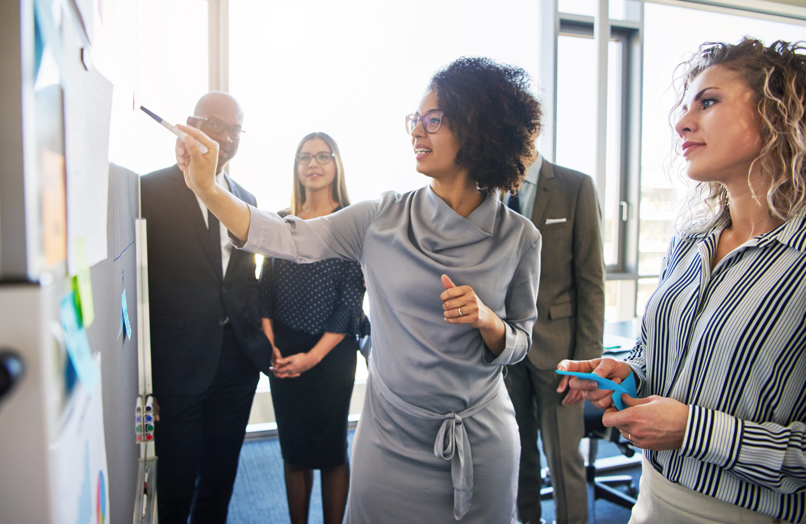 Diverse group of focused business people brainstorming together on a whiteboard during a strategy session in a bright, modern office.