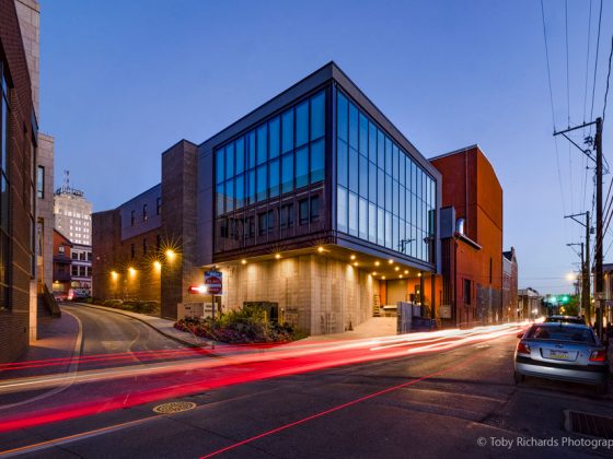 A historic theatre with tall glass windows looking into a dance studio in the evening with cars passing quickly on the street.