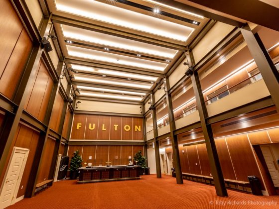 A brown wood paneled lobby with a front desk, renovated in the historic theatre. Large letters on the wall spell out Fulton.