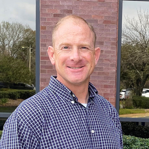 A Caucasian man in blue plaid button-down shirt smiles outdoors for a picture.