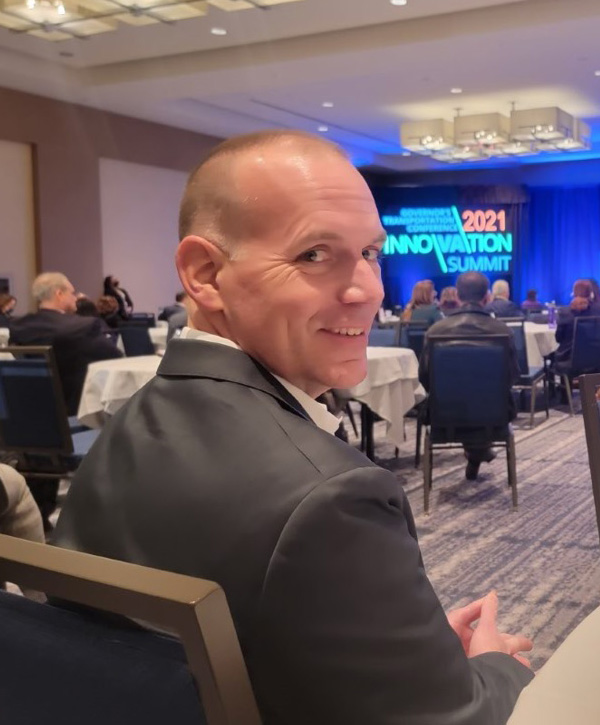 Bryan is sitting at a table at the 2021 Innovation Summit. He is facing forward but is turned around to look at the camera and is smiling.