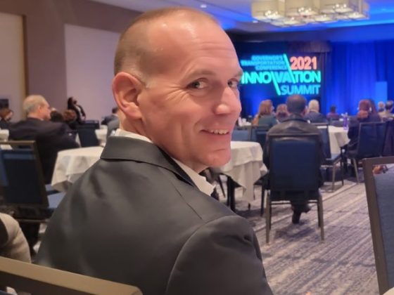 Bryan is sitting at a table at the 2021 Innovation Summit. He is facing forward but is turned around to look at the camera and is smiling.