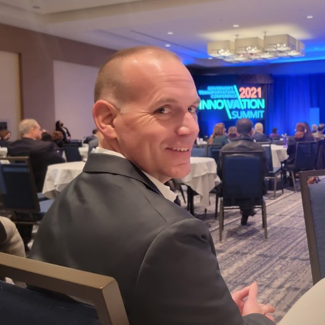 Bryan is sitting at a table at the 2021 Innovation Summit. He is facing forward but is turned around to look at the camera and is smiling.