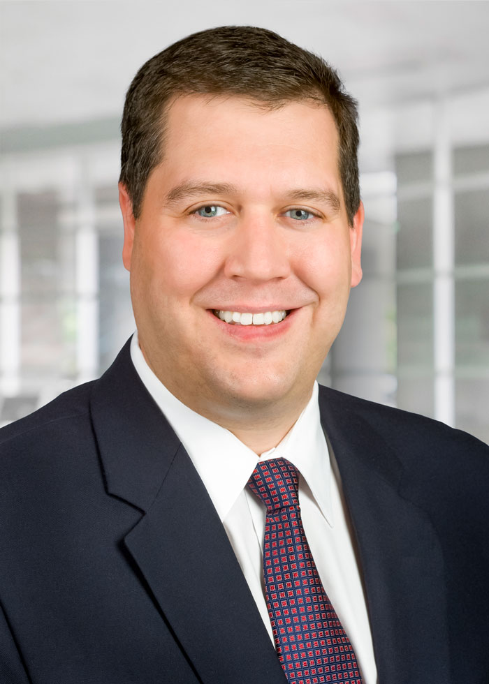 A man with brown hair in a white shirt with a red and blue tie and black suit jacket smiling for the camera.