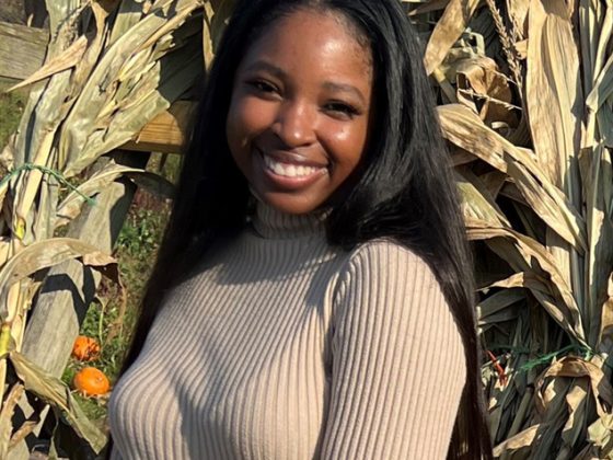 Construction Services Project Manager Emani Majors stands outside smiling in front of some corn stalks on a sunny day while wearing white shorts and a cream-colored long-sleeve shirt.