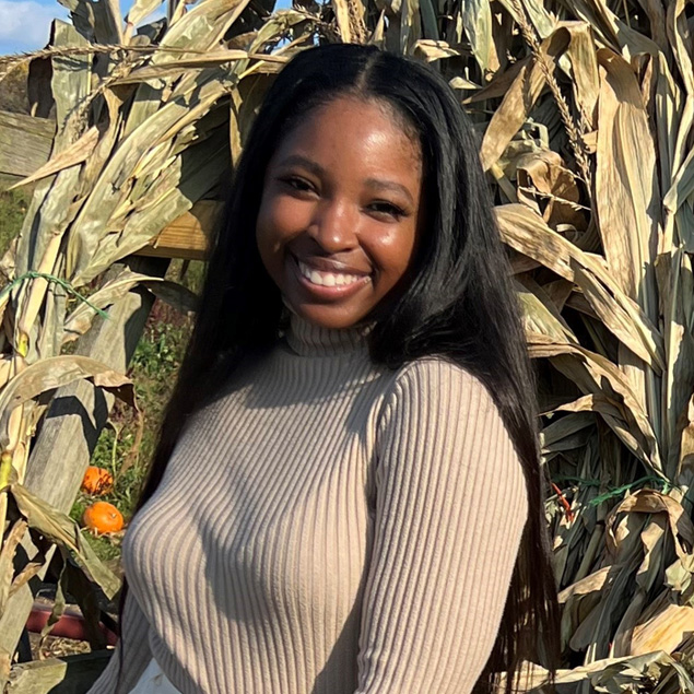 Construction Services Project Manager Emani Majors stands outside smiling in front of some corn stalks on a sunny day while wearing white shorts and a cream-colored long-sleeve shirt.