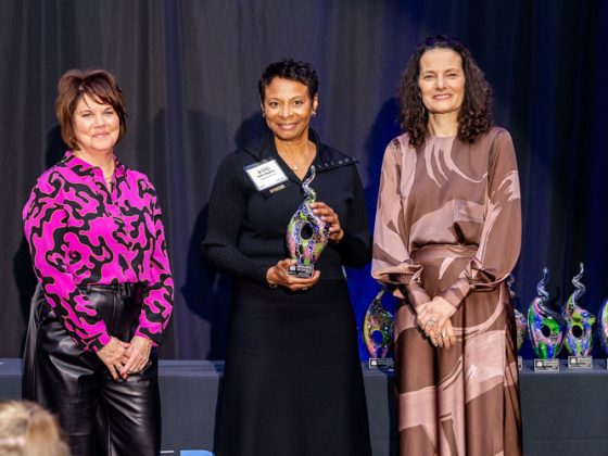 Three women stand smiling on a stage with a black curtain behind them. The woman in the center is holding a glass award.