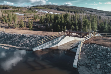 Water flows under the pedestrian bridge of a dam’s concrete chute spillway with pine tree-covered hills in the background.