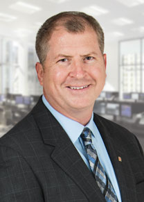 George Olear wearing a dark suit jacket with a light blue shirt and blue and black striped tie smiling for the camera.