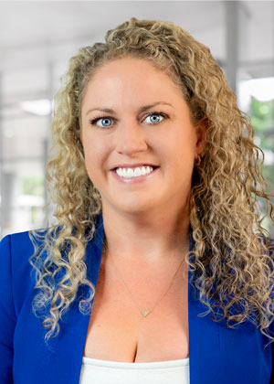 A woman is posing for a professional headshot. She is wearing a white shirt and royal blue jacket. She is standing in an office lobby.