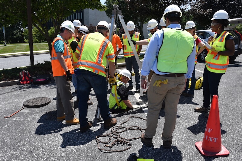 Gannett Fleming employees in high visibility vests and hard hats watch as their coworker descends into a manhole.