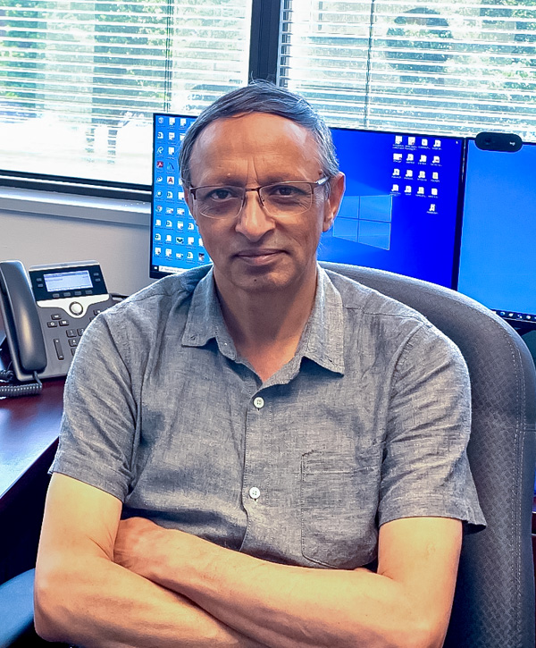 Qasir sits with his arms folded in an office chair in front of two large windows with open blinds. There are two computer monitors on his desk.