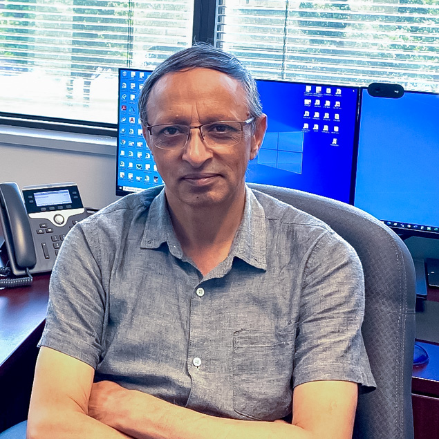 Qasir sits with his arms folded in an office chair in front of two large windows with open blinds. There are two computer monitors on his desk.