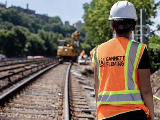 A female construction professional wearing a safety jacket and hard hat overlooks a railroad.