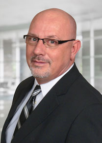 A male wearing glasses in a dark suite with a white shirt and blue stripped tie smiling.