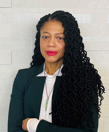 A black women in a dark suit with a white shirt standing in front of a white brick wall.