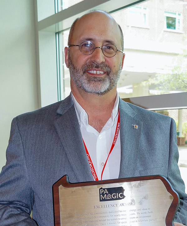 Dave Gilbert receives an Excellence Award from PA MAGIC. Wearing a grey suit jacket, he stands in front of a window holding the award in front of him.