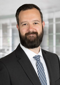 A man poses for a professional headshot. He is smiling and wearing a dark gray sports coat, white shirt, and blue striped tie.