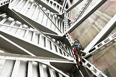 A person wearing a harness and hard hat uses a rope to climb a metallic and concrete structure.