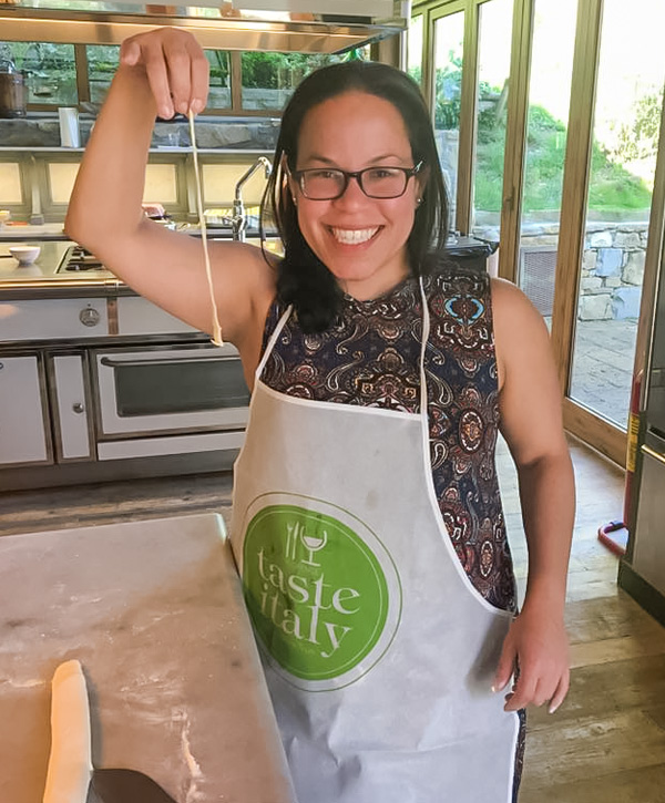 Circe wearing a whie apron taking a cooking lesson holding up a hand-rolled noodle smiling for the camera.